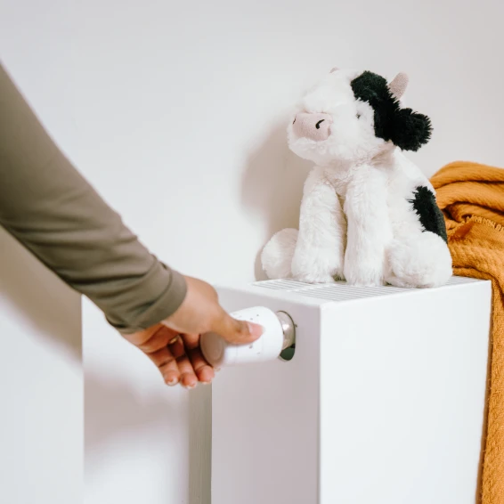 Hand draait aan de radiatorknop van een verwarming, waar een knuffel van een koe op staat.
