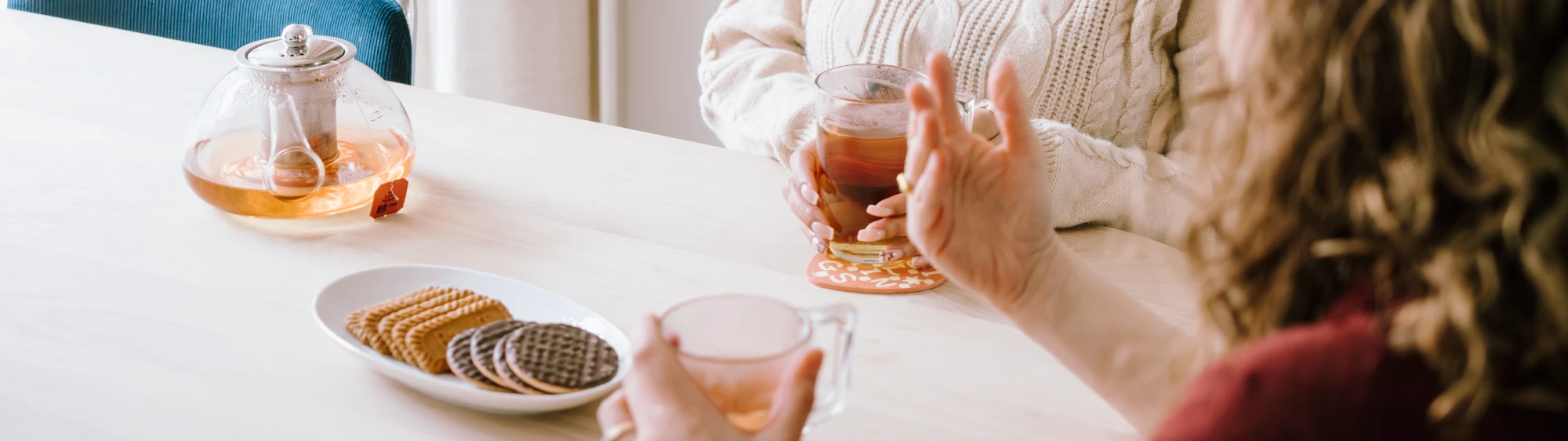Op de foto zie je twee paar handen van twee vrouwen die aan tafel zitten en thee drinken. Er staan ook koekjes op tafel.
