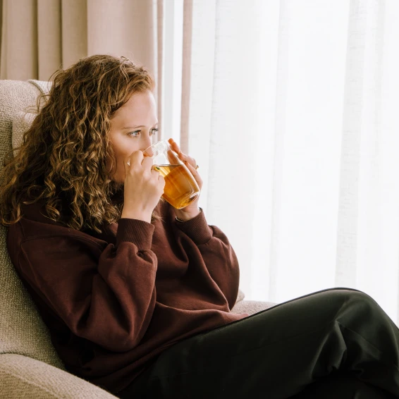 Op de foto zit een jonge vrouw met krullend haar op een comfortabele stoel thee te drinken.