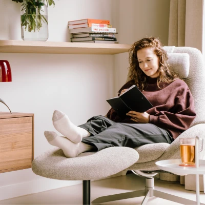 Op de foto zit een jonge vrouw met krullend haar een boek te lezen in een comfortabele stoel.