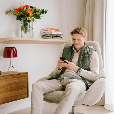 Op de foto zit een jonge man met een groen vest over een witte trui in een comfortabele beige stoel op zijn telefoon iets te lezen. Naast hem hangt een plank aan de muur met daarop bloemen in een vaas en een rode lamp.