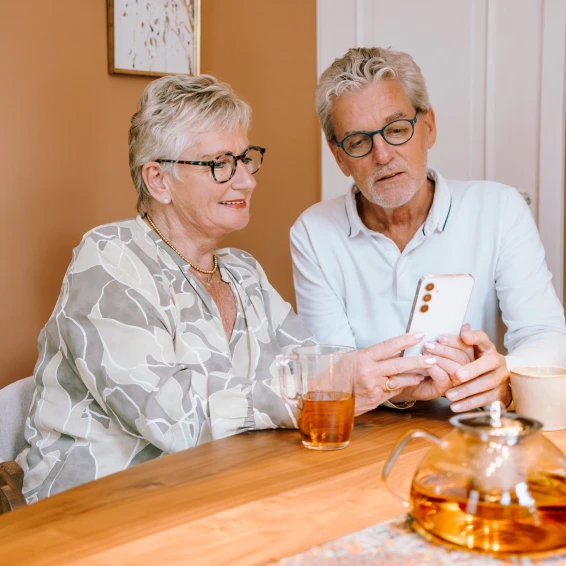 Op de foto staat een ouder echtpaar. Ze zitten aan tafel en lezen samen wat op een telefoon.